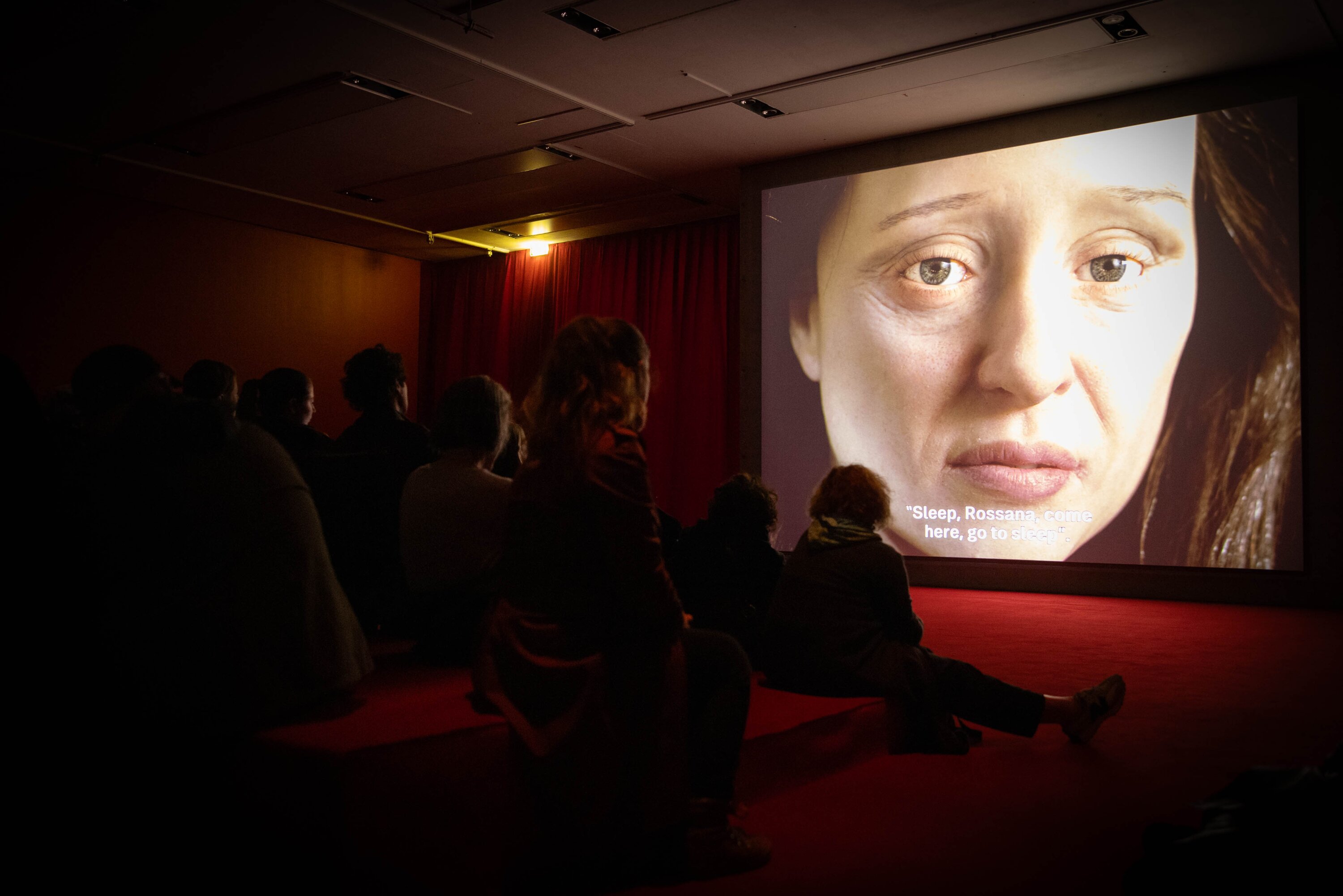 Personen sitzen in einem dunklen Raum und schauen auf eine Leinwand, die das Gesicht einer Frau in Gro&szlig;aufnahme zeigt.