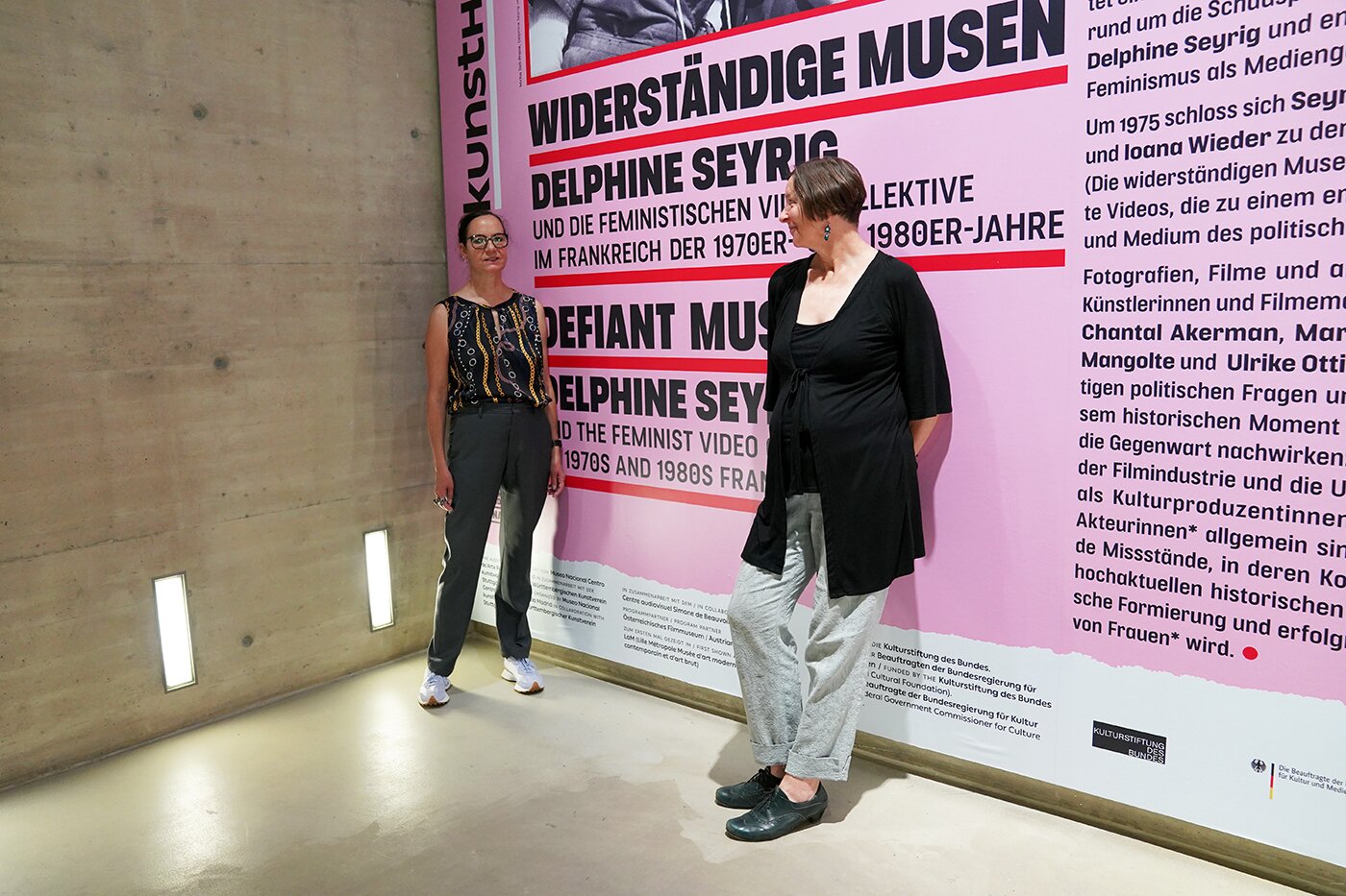 Zwei Frauen stehen vor der Plakatwand der Ausstellung Widerständige Musen