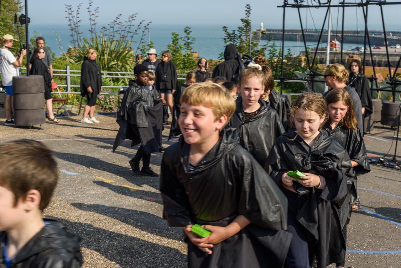 Kinder in schwarzen Regencapes laufen im Kreis vor einer Küstenlandschaft.