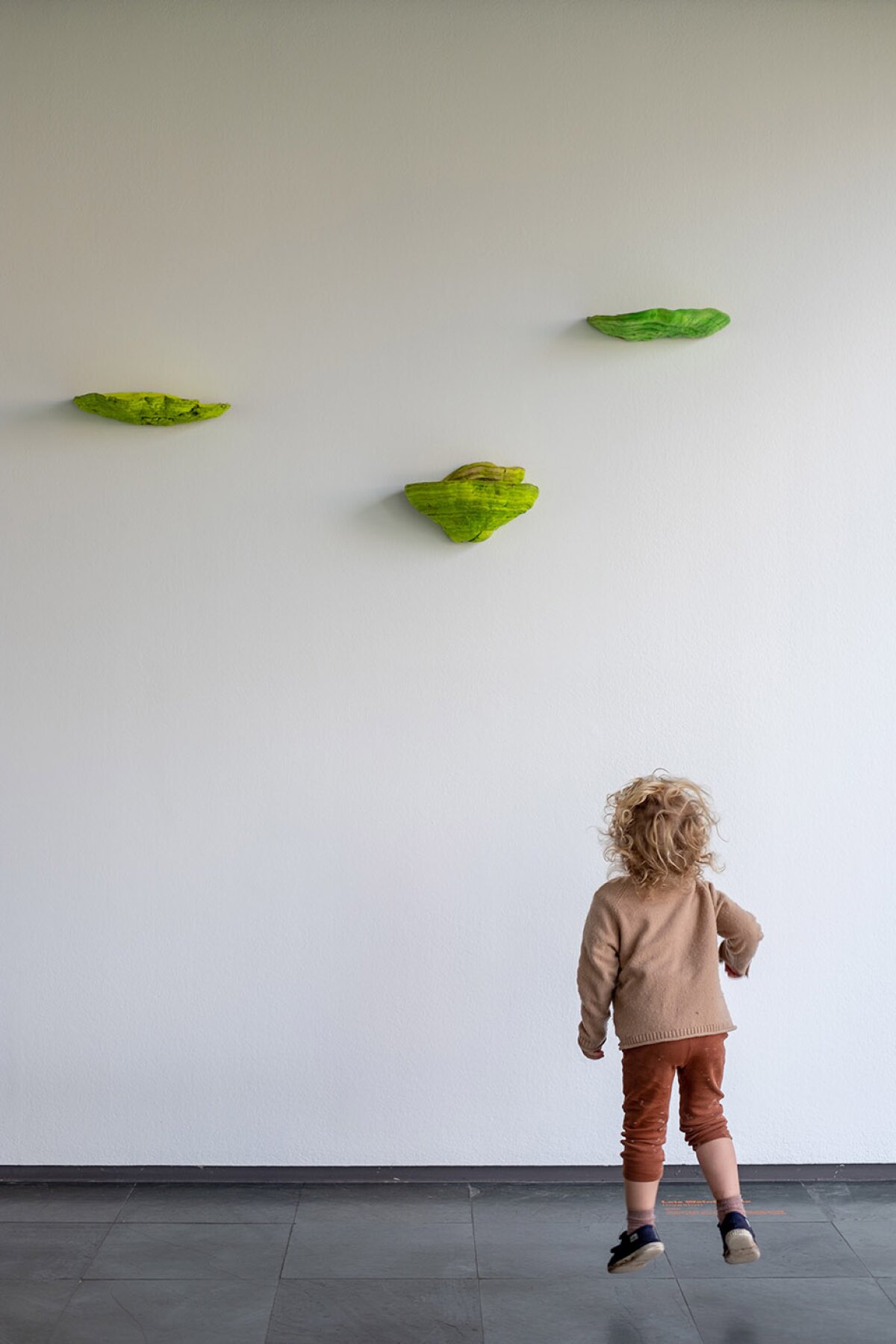 Ausstellungsansicht, kleines Kind h&uuml;pft vor wei&szlig;er Wand mit gr&uuml;nen Pilzen