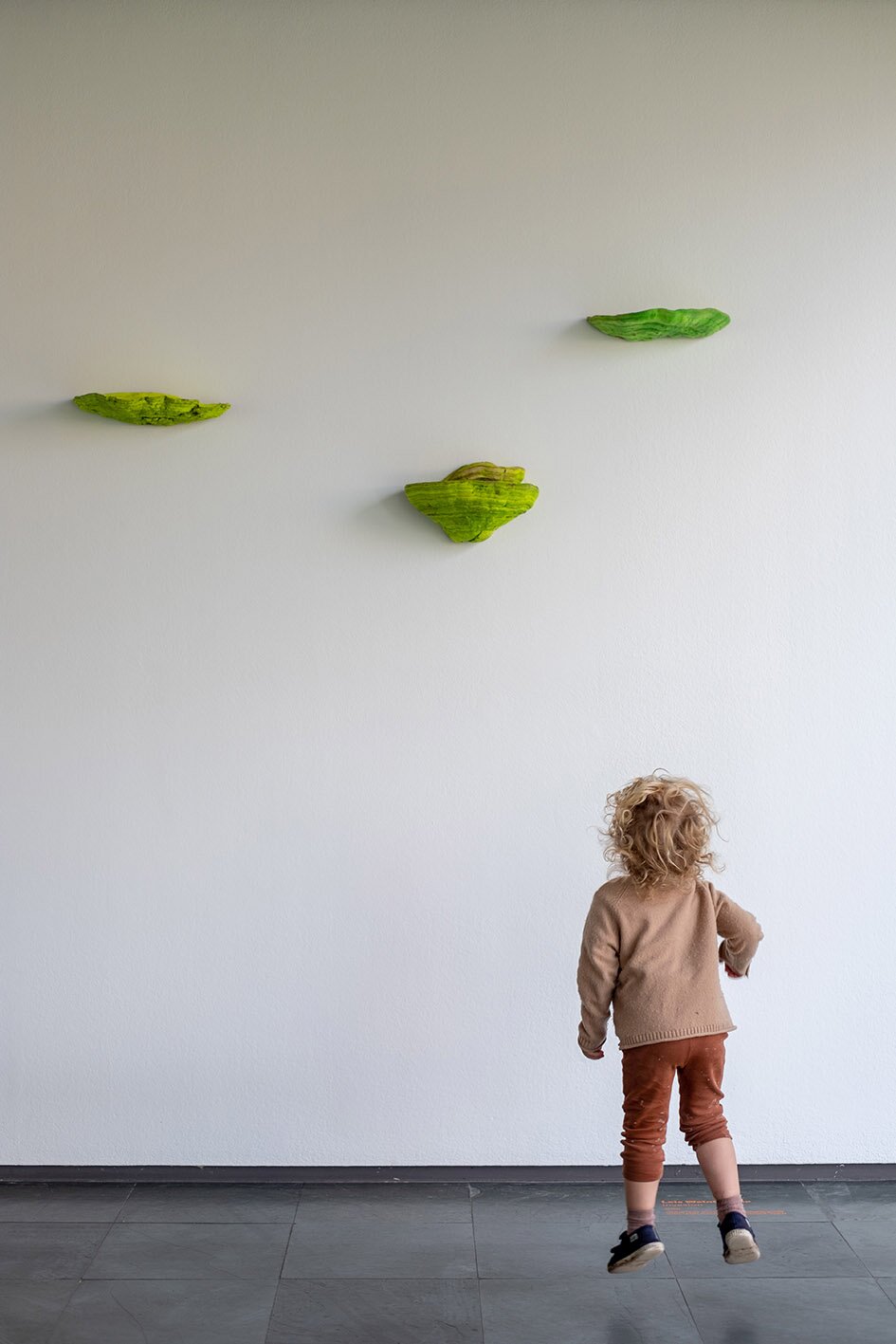 Ausstellungsansicht, kleines Kind hüpft vor weißer Wand mit grünen Pilzen