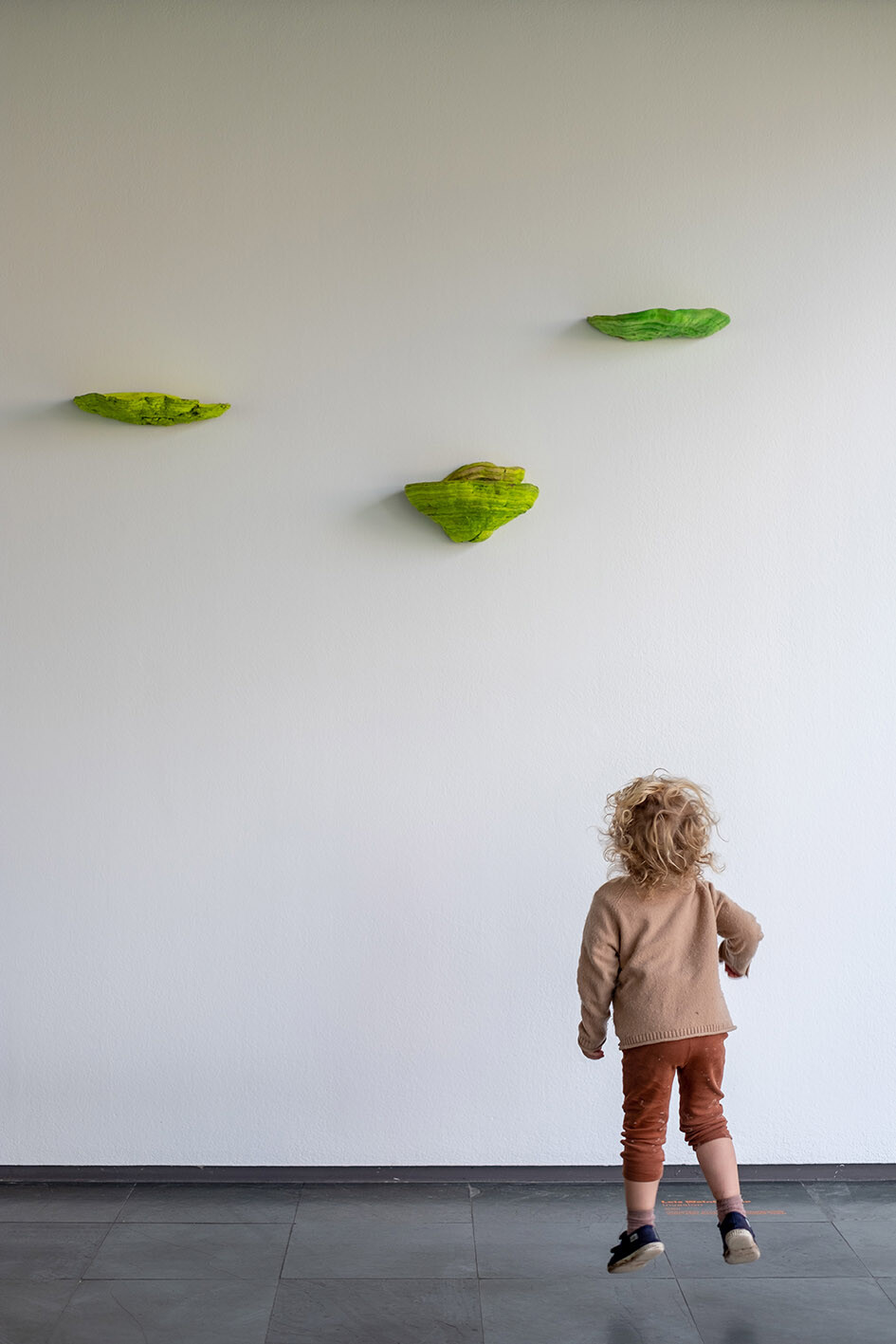 Ausstellungsansicht, kleines Kind hüpft vor weißer Wand mit grünen Pilzen