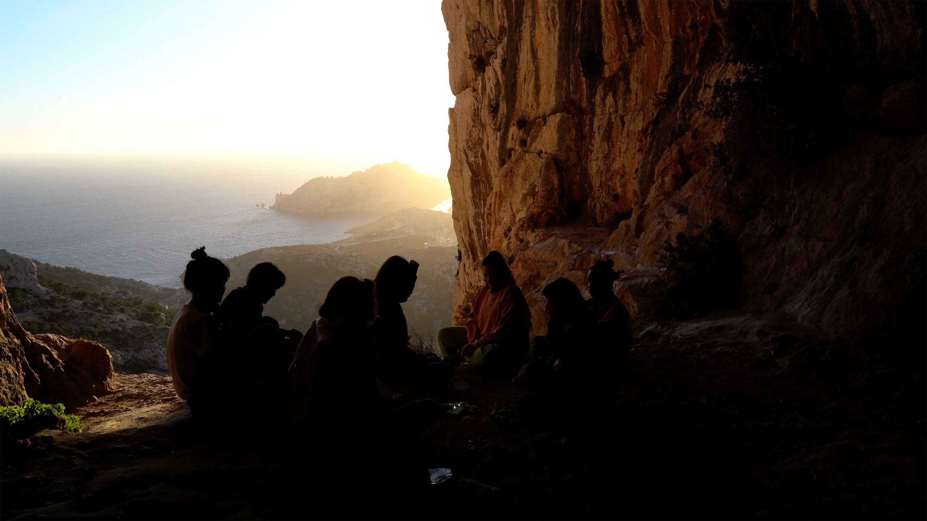 Eine Gruppe Menschen sitzt in einer Höhle, im Hintergrund ist das Meer.