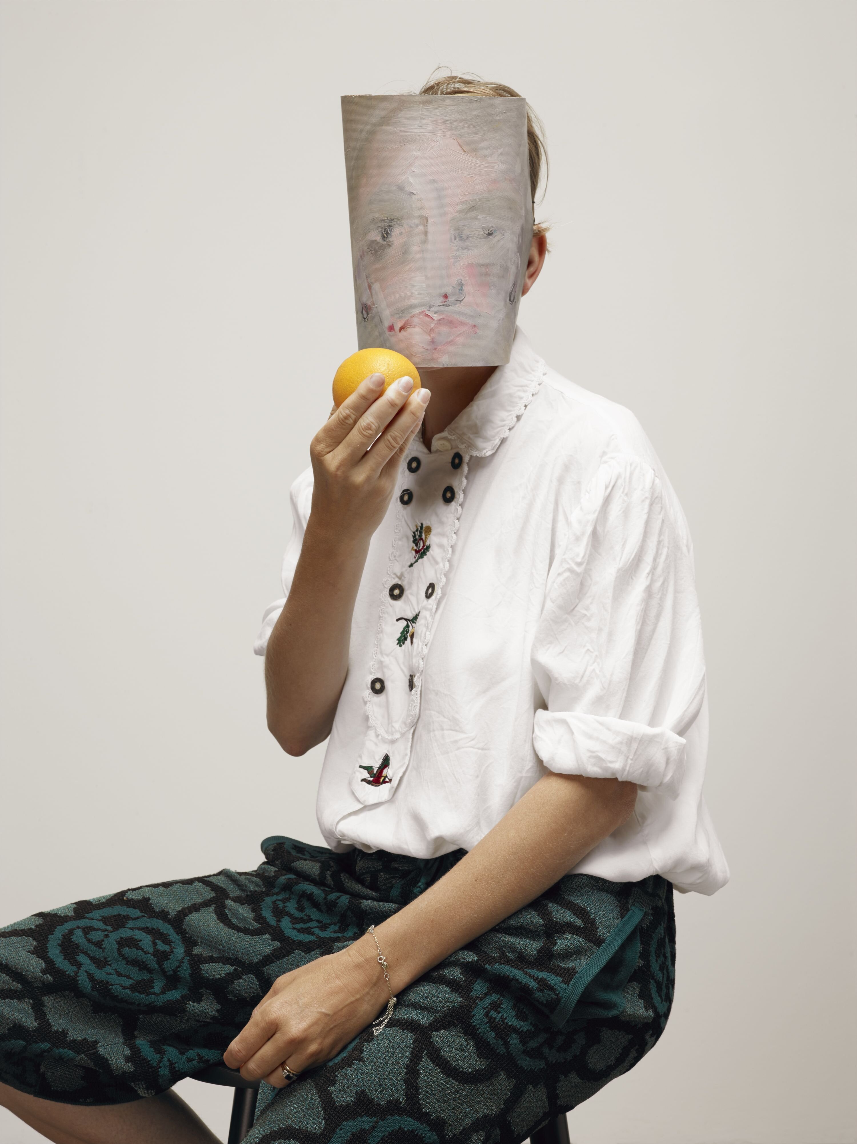 Person mit einer Maske eines gemalten Gesichtes sitz und hat einen Apfel in der Hand