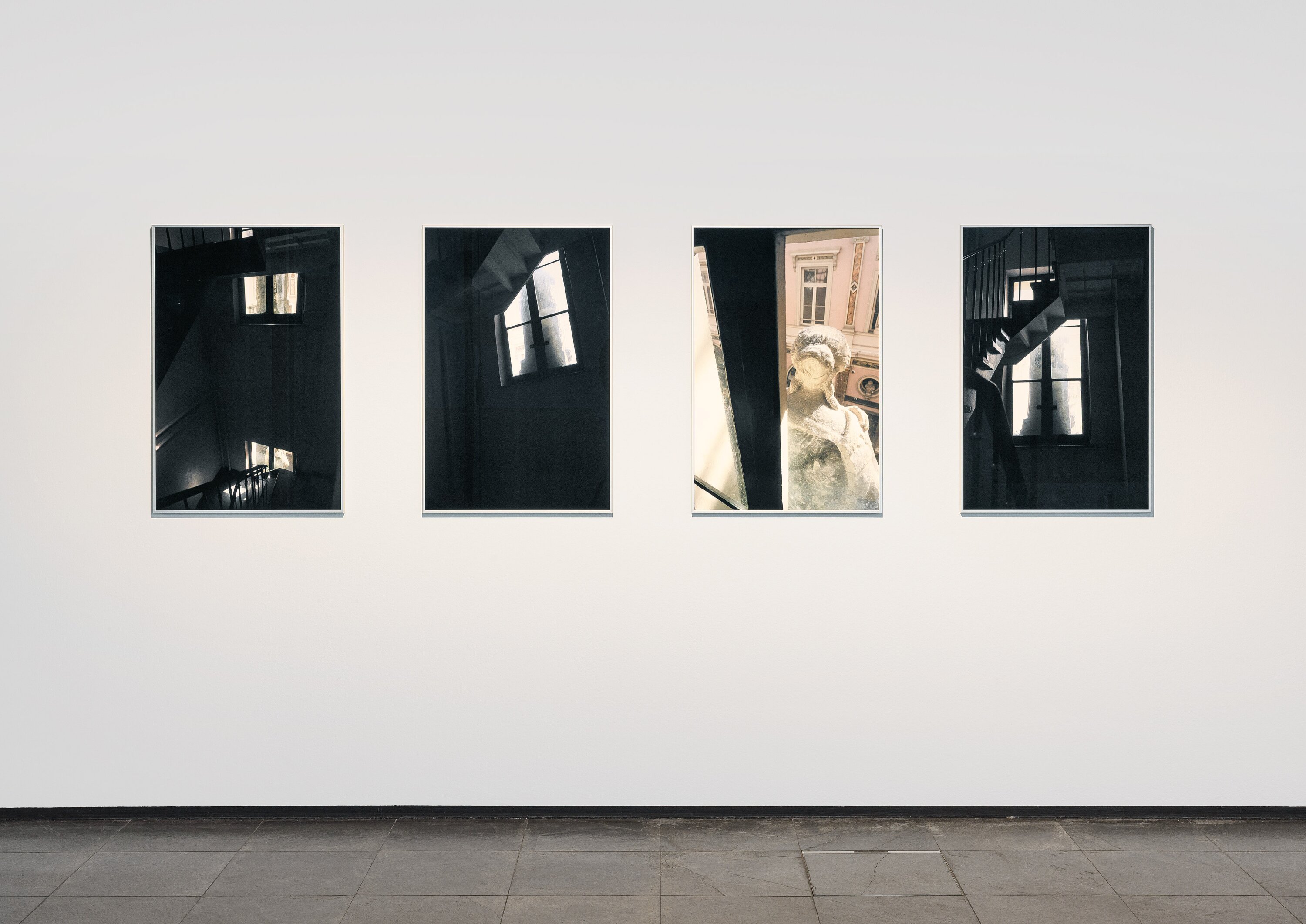 Ausstellungsansicht mit vier Fotografien, die ein Treppenhaus und eine Statur aus verschiedenen Perspektiven zeigen.